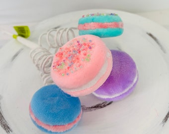 May include: Three pastel-colored macarons with white filling. The macarons are decorated with sprinkles and are arranged on a white surface.