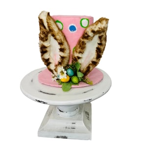 以下が含まれることがあります： ピンクのイースターバニーハットは、茶色の毛皮の耳、緑と青のドット、緑の葉と青と黄色のイースターエッグのフラワーアレンジメントが付いています。ハットは白い台座の上に置かれています。