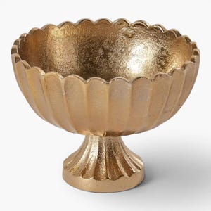 May include: A gold-coloured, pedestal bowl with a scalloped rim. The bowl has a textured surface and a rounded base. The bowl is made of metal and is likely used for decorative purposes or to hold small items.