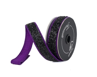 Peut inclure: Une bobine de ruban décoratif avec un bord noir perlé et un dos en tissu violet foncé. Le ruban est partiellement déroulé, révélant les couleurs et les textures contrastées. La bobine est noire avec du texte blanc.
