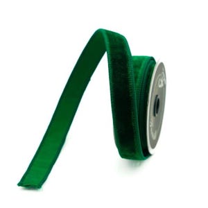 Può includere: Una bobina di nastro in velluto verde smeraldo intenso con una consistenza morbida e vellutata. Il nastro è avvolto attorno a un nucleo circolare in plastica nera, con un'estremità che si estende verso l'esterno per mostrare il colore uniforme.