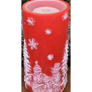 May include: A red Christmas candle with white snowflake and snowman designs. The candle has a cylindrical shape and a white interior. The base of the candle features a detailed relief of snowmen and fir trees.