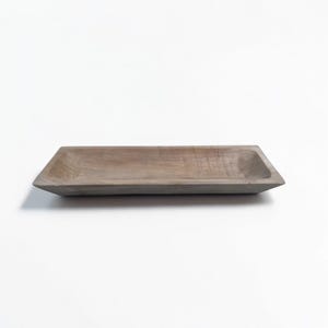 May include: A rectangular wooden tray with a light brown finish. The tray has a shallow, open design with angled sides and a flat bottom. The wood grain is visible, adding texture to the piece. Ideal for serving or display.