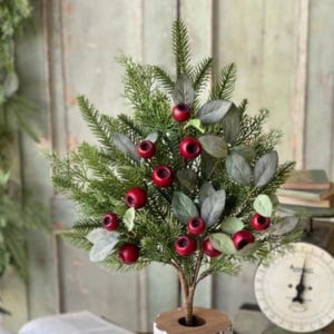 May include: A decorative artificial plant featuring dense green pine needles and sage-colored leaves accented with small, round, deep red berries. The plant is supported by a central brown stem that extends into a circular wooden base.