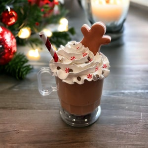 Könnte beinhalten: Ein klarer Becher gefüllt mit einem schokoladenfarbenen Getränk, garniert mit Schlagsahne, Schokoladenstückchen und Pfefferminzbonbons. Ein Lebkuchenmann-Keks und ein rot-weiß gestreifter Strohhalm sind enthalten. Festliches Urlaubsthema.