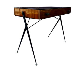 以下が含まれることがあります： 1つの引き出しと黒い金属製の脚が付いた、ミッドセンチュリーモダンな机。机の天板は、薄い茶色の仕上げの木材で作られています。