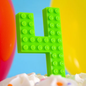 May include: A green number four birthday candle made of plastic LEGO bricks. The candle is sitting on a white cake with colorful sprinkles.