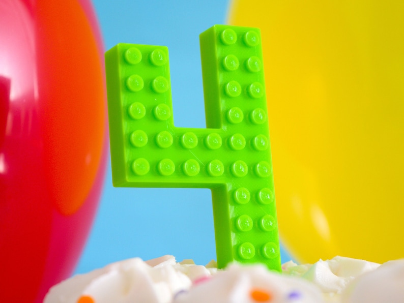 May include: A green number four birthday candle made of plastic LEGO bricks. The candle is sitting on a white cake with colorful sprinkles.