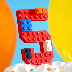 May include: A red Lego number five cake topper with blue, white, and orange Lego bricks. The number five is sitting on a white frosted cake with colorful sprinkles.