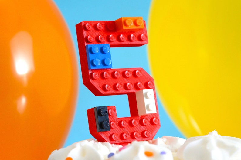 May include: A red Lego number five cake topper with blue, white, and orange Lego bricks. The number five is sitting on a white frosted cake with colorful sprinkles.