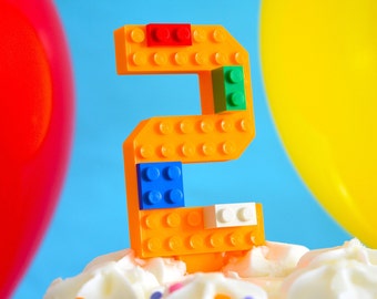May include: A bright orange number two made of Lego blocks, with red, blue, green, and white accents, sits on top of a white frosted cake.