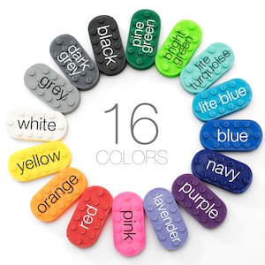 May include: A circle of 16 small, rectangular plastic pieces, each a different color, with the color name printed on each piece. The colors are: black, dark grey, grey, white, yellow, orange, red, pink, lavender, purple, navy, blue, light blue, turquoise, light green, and pine green. The text "16 COLORS" is printed in the center of the circle.