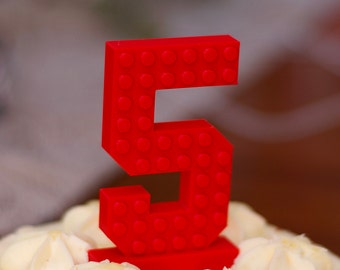 May include: A red plastic number five shaped like a Lego brick, sitting on top of a white cake with white frosting.