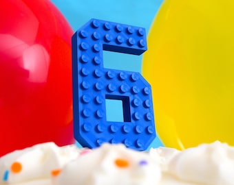 May include: A blue plastic number six shaped like a Lego brick. The number is sitting on top of a white cake with colorful sprinkles.