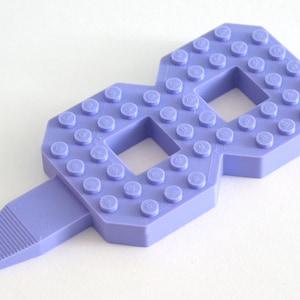 May include: A purple plastic number eight made from Lego bricks. The number eight has a small handle on the bottom.