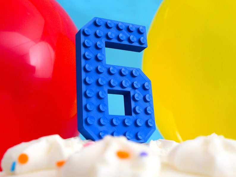 May include: A blue plastic number six shaped like a Lego brick. The number is on top of a white cake with colorful sprinkles.