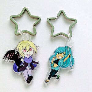 May include: Two character charms with star-shaped keychains. The charm on the left features a blonde character with a black bat wing and a purple scarf. The charm on the right features a blue-haired character with a white staff.