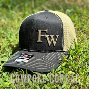 May include: Black and beige trucker hat with a gold embroidered "FW" logo. The hat is made by Richardson 112.