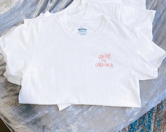 May include: White t-shirt with the text "COFFEE TIL COCKTAILS" embroidered in pink and orange on the front.