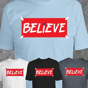 May include: A light blue t-shirt with a red rectangle featuring the word "BELIEVE" in white, and a blue star. Below are white, black, and grey t-shirts with the same design.