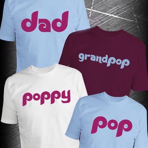 May include: Four t-shirts with text in a playful font. One light blue shirt says "dad" in pink, a burgundy shirt says "grandpop" in light blue, a white shirt says "poppy" in pink, and a light blue shirt says "pop" in pink.