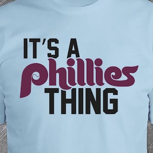 May include: A light blue t-shirt with the text "It's a Phillies Thing" in black and maroon lettering.