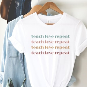 May include: White t-shirt with the words "teach love repeat" printed in a repeating pattern of pastel colors. The shirt is hanging on a wooden hanger with a denim shirt and a brown hat in the background.