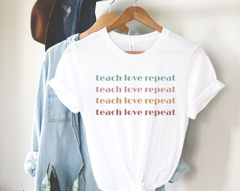 May include: White t-shirt with the words "teach love repeat" printed in a repeating pattern of pastel colors. The shirt is hanging on a wooden hanger with a denim shirt and a brown hat in the background.