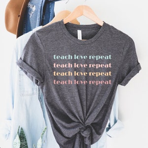 May include: Dark gray t-shirt with the words "teach love repeat" printed in pastel colors. The shirt is hanging on a wooden hanger with other clothing items in the background. The shirt has rolled-up sleeves.