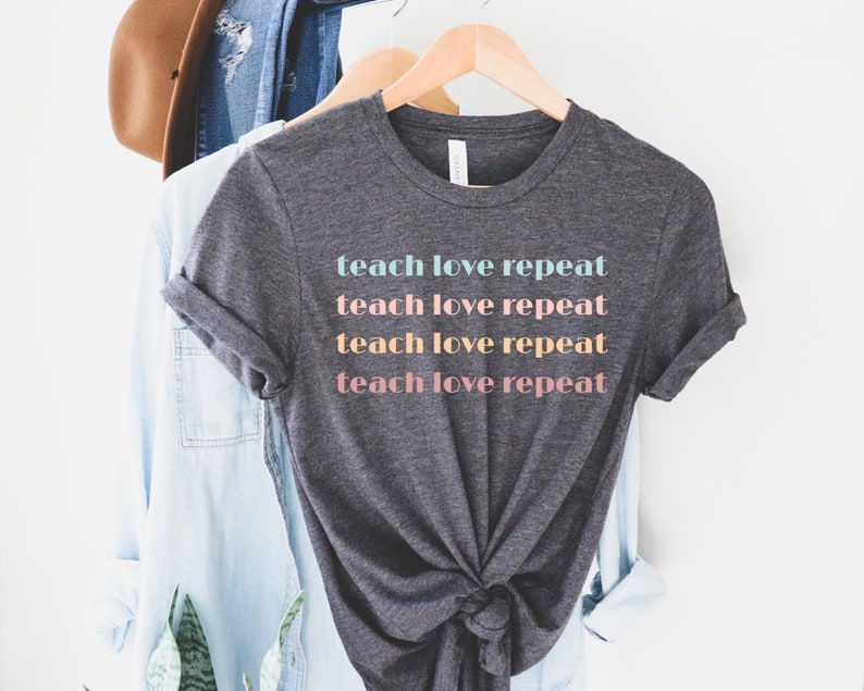 May include: Dark gray t-shirt with the words "teach love repeat" printed in pastel colors. The shirt is hanging on a wooden hanger with other clothing items in the background. The shirt has rolled-up sleeves.