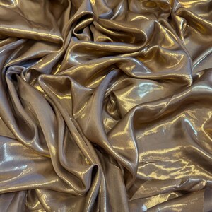 Op de afbeelding: Close-up van een vloeiende, glinsterende stof in een warme, goud-bronzen tint. Het materiaal is sterk gedrapeerd, waardoor diepe plooien en highlights ontstaan die het licht vangen. De algehele textuur lijkt zacht en luxueus, wat duidt op een hoogwaardig textiel.