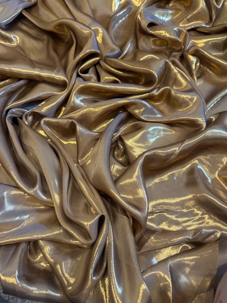 Op de afbeelding: Close-up van een vloeiende, glinsterende stof in een warme, goud-bronzen tint. Het materiaal is sterk gedrapeerd, waardoor diepe plooien en highlights ontstaan die het licht vangen. De algehele textuur lijkt zacht en luxueus, wat duidt op een hoogwaardig textiel.