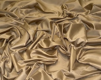 Può includere: Primo piano di un grande pezzo di tessuto con molte pieghe. Il tessuto è di un colore marrone dorato caldo e uniforme con una leggera lucentezza, creando riflessi e ombre. Il materiale sembra morbido e fluido, forse seta o un tessuto simile.