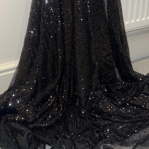 Può includere: Un lungo abito nero con corpetto di paillettes e gonna fluida. Il tessuto brilla con paillettes sparse e l'orlo è rifinito con un tessuto trasparente a balze. L'abito sembra essere realizzato in un materiale morbido e drappeggiato.