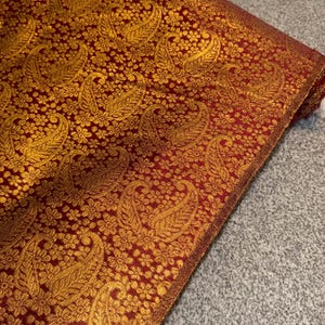 May include: A folded length of fabric with an elaborate design. The material has a deep red base, overlaid with a repeating pattern of gold paisley shapes and floral details. The edges are finished with a gold border.
