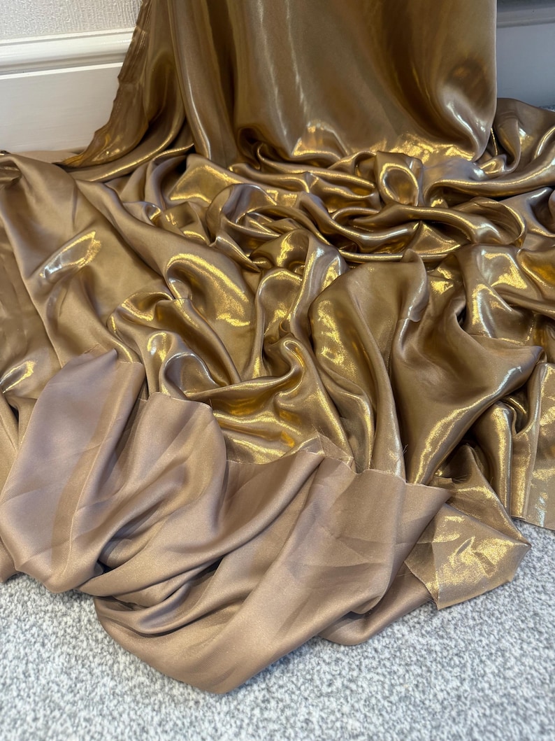 Op de afbeelding: Een luxueuze, vloeiende stof in goud- en bronskleuren. Het materiaal is gedrapeerd en geplooid, waardoor zachte plooien en highlights ontstaan. De glanzende, satijnachtige textuur reflecteert het licht, wat diepte en dimensie aan de stof toevoegt.