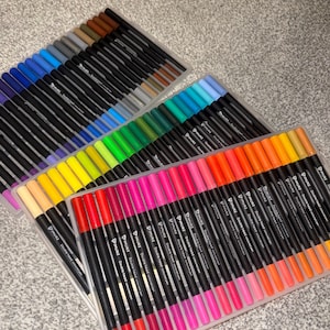 May include: A selection of double-ended coloured pens in clear plastic cases, arranged to display a variety of colours. The pens are in shades of blue, green, yellow, pink, and orange. Each pen has a black barrel with the brand name printed on it.