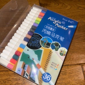 May include: A set of 36 acrylic markers in a clear plastic case. The packaging is blue and white, with the words "Acrylic Marker" and Chinese characters. The markers are white with colored tips, and the packaging shows examples of artwork.