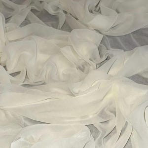 Può includere: Tessuto bianco sporco e fluente con un aspetto morbido e traslucido. Il materiale è drappeggiato e raccolto, creando un effetto a strati con ombre e luci sottili. Il tessuto appare leggero e arioso, adatto per abbigliamento o scopi decorativi.