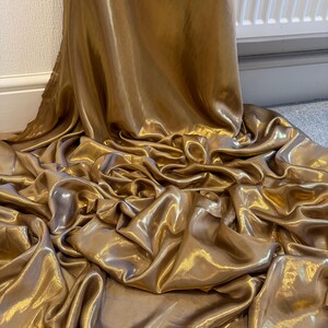 Op de afbeelding: Close-up van een vloeiende, gouden stof met een glanzende glans. Het materiaal is gedrapeerd en geplooid, waardoor diepe plooien en schaduwen ontstaan. De kleur van de stof is een rijk, warm goud, dat licht reflecteert en een luxueus gevoel geeft.