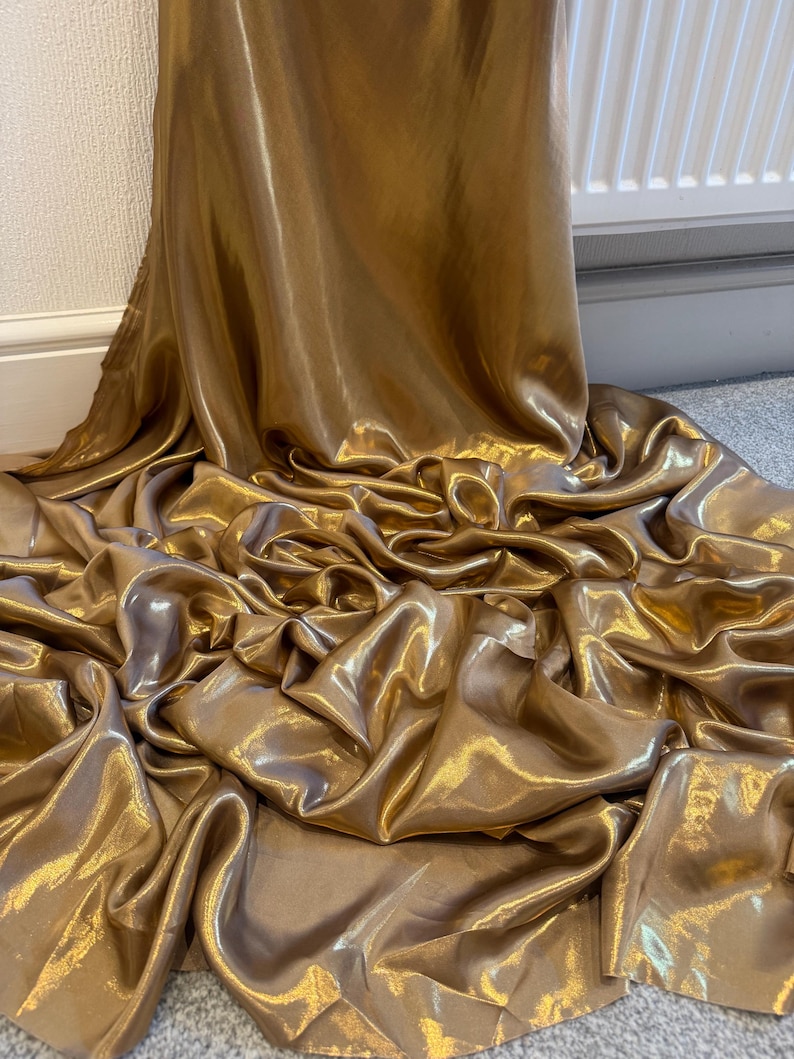 Op de afbeelding: Close-up van een vloeiende, gouden stof met een glanzende glans. Het materiaal is gedrapeerd en geplooid, waardoor diepe plooien en schaduwen ontstaan. De kleur van de stof is een rijk, warm goud, dat licht reflecteert en een luxueus gevoel geeft.