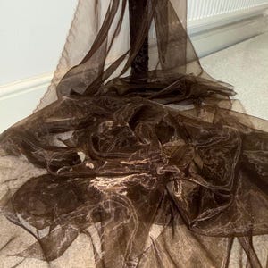 Peut inclure: Un tissu transparent brun foncé drapé et froissé. Le matériau présente un aspect chatoyant, la lumière se reflétant sur sa surface. Le tissu semble léger et est probablement destiné à un usage décoratif, comme des rideaux ou des tentures.