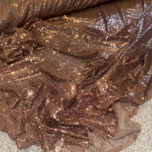 Peut inclure: Un rouleau de tissu à paillettes marron. Le tissu est orné de petites paillettes réfléchissantes qui captent la lumière, créant un effet scintillant. Le matériau est drapé et plié, mettant en valeur sa texture et son tombé.