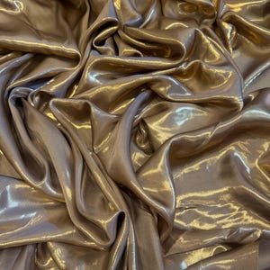 Op de afbeelding: Close-up van een luxueuze, vloeiende stof in een warme, goud-bronzen tint. Het materiaal is gedrapeerd en gevouwen, waardoor een spel van licht en schaduw over het glanzende oppervlak ontstaat. De stof lijkt zacht en vloeiend.