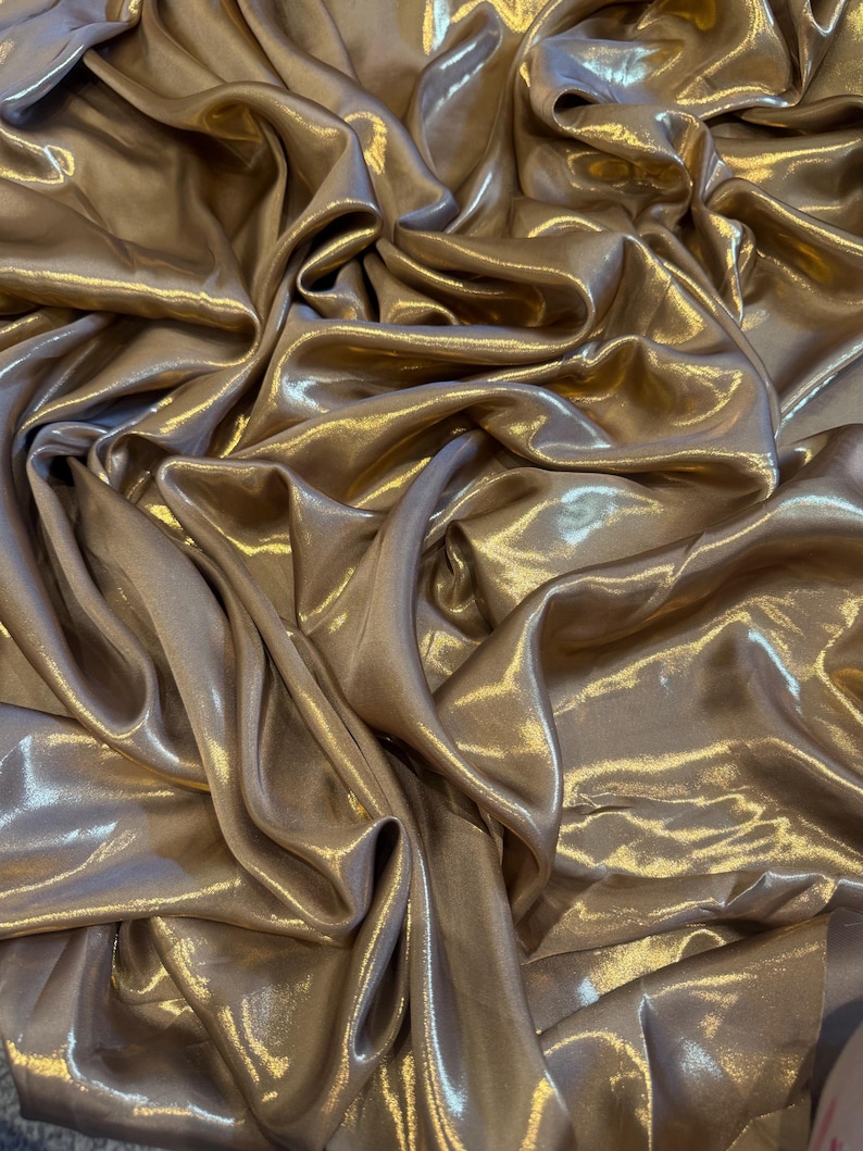 Op de afbeelding: Close-up van een luxueuze, vloeiende stof in een warme, goud-bronzen tint. Het materiaal is gedrapeerd en gevouwen, waardoor een spel van licht en schaduw over het glanzende oppervlak ontstaat. De stof lijkt zacht en vloeiend.