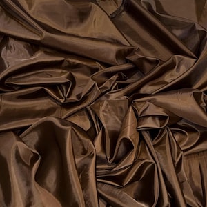 May include: A length of dark brown fabric with a smooth, slightly reflective surface. The material is gathered and draped in multiple folds, creating deep shadows and highlights across the surface, showcasing its texture and deep brown colour.