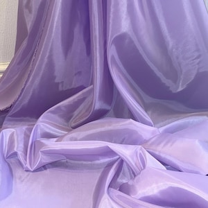 May include: Close-up of a flowing, light purple fabric. The material has a shiny, silky texture with soft folds and highlights. The fabric appears lightweight and drapes gracefully, suggesting it could be used for clothing or decorative purposes.