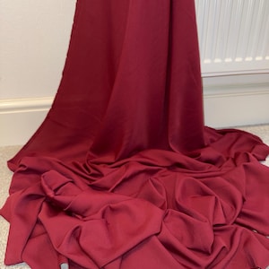 May include: A length of solid deep burgundy fabric with a smooth, matte texture. The material is draped in soft, flowing folds that cascade downward, creating a series of gentle curves and shadows across the surface of the textile.
