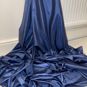 Könnte beinhalten: Ein großer Bereich aus marineblauem Stoff, möglicherweise ein Vorhang oder eine Draperie, ist gerafft und drapiert. Das Material hat ein glänzendes, satinartiges Aussehen, mit Falten und Schatten, die Tiefe und Textur erzeugen. Der Stoff liegt auf einer hellen Oberfläche.