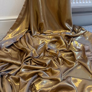 Op de afbeelding: Een luxueuze, goudkleurige stof met een glanzende satijnen afwerking. Het materiaal is gedrapeerd en gevouwen, wat een gevoel van beweging en luxe cre&euml;ert. De kleur is een warm, rijk goud, en de stof lijkt uit &eacute;&eacute;n stuk te bestaan.